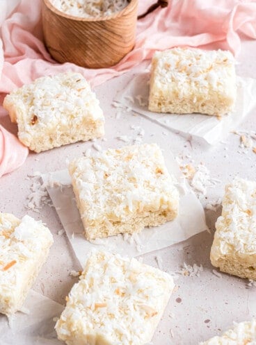 Coconut bar squares on pieces of parchment paper in front of a pink linen napkin and a wooden bowl with coconut.