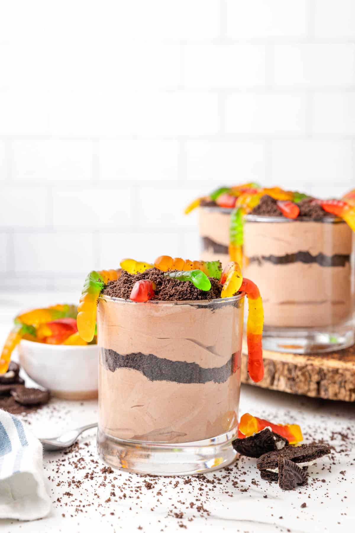 An individual dirt cup with more desserts and gummy worms in the background.