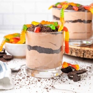 Cups of dirt with gummy worms with crumbs and candy around them.