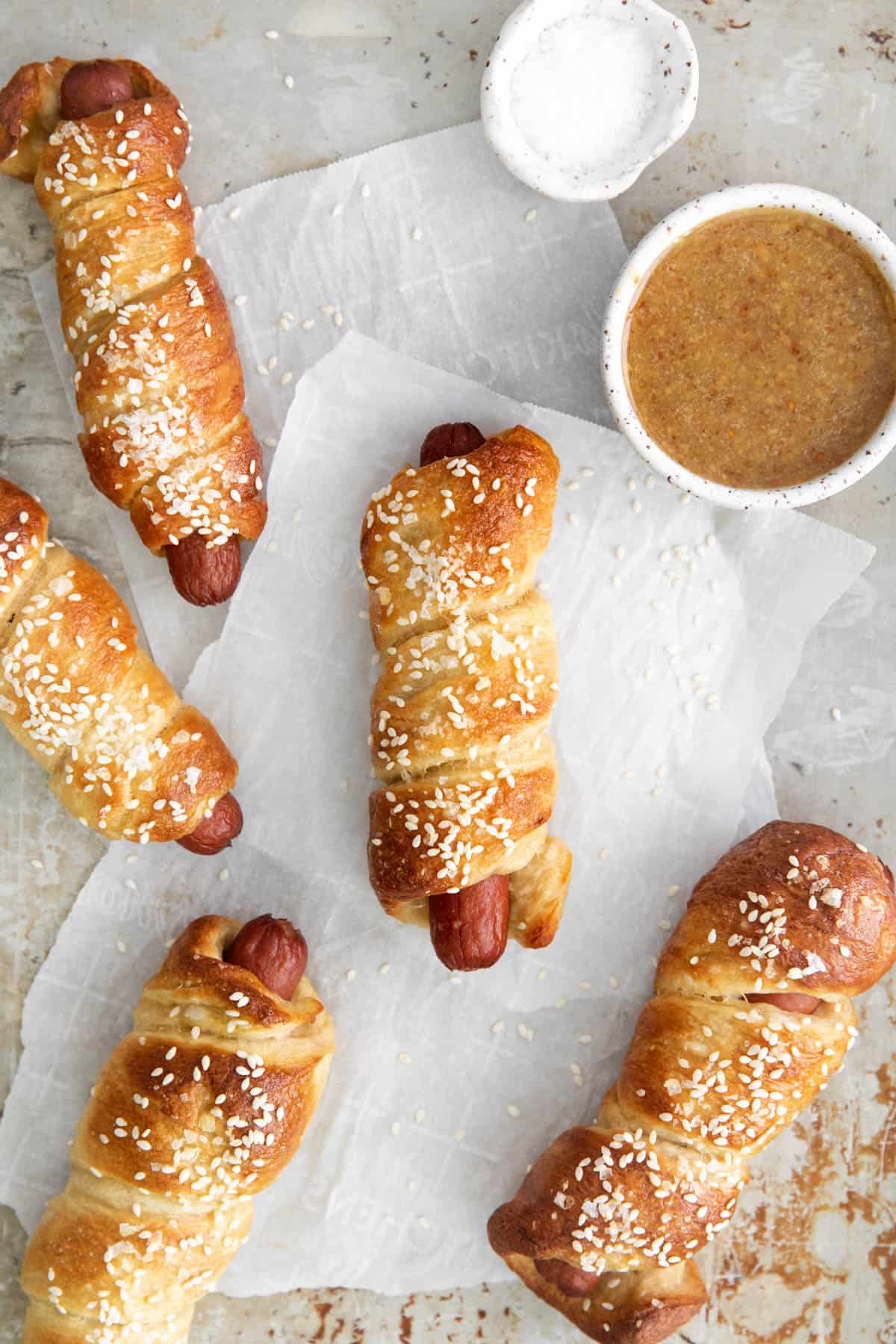 Soft Pretzel Dogs on parchment paper next to bowls of dipping sauce and salt.