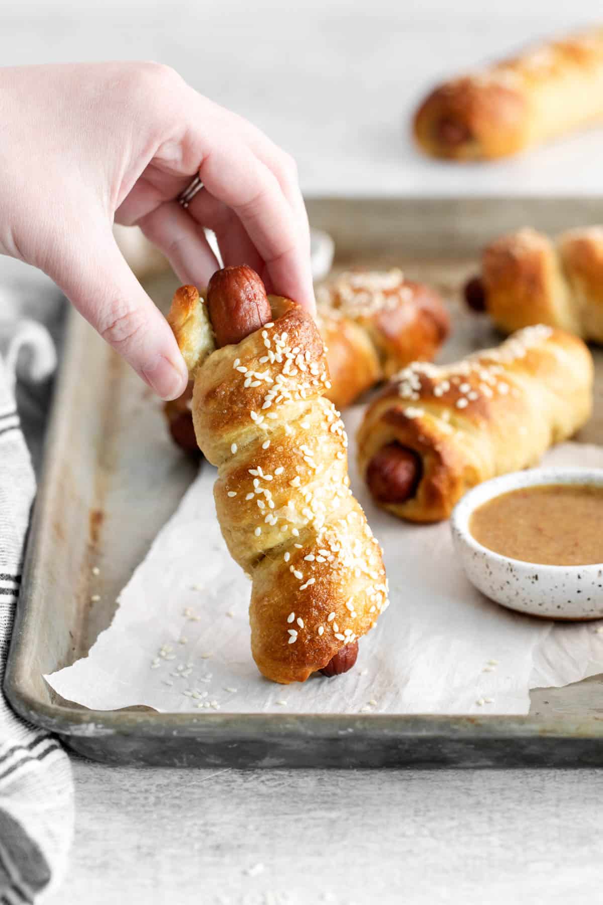 A hand holding a soft pretzel dog on a baking sheet with more pretzel dogs.
