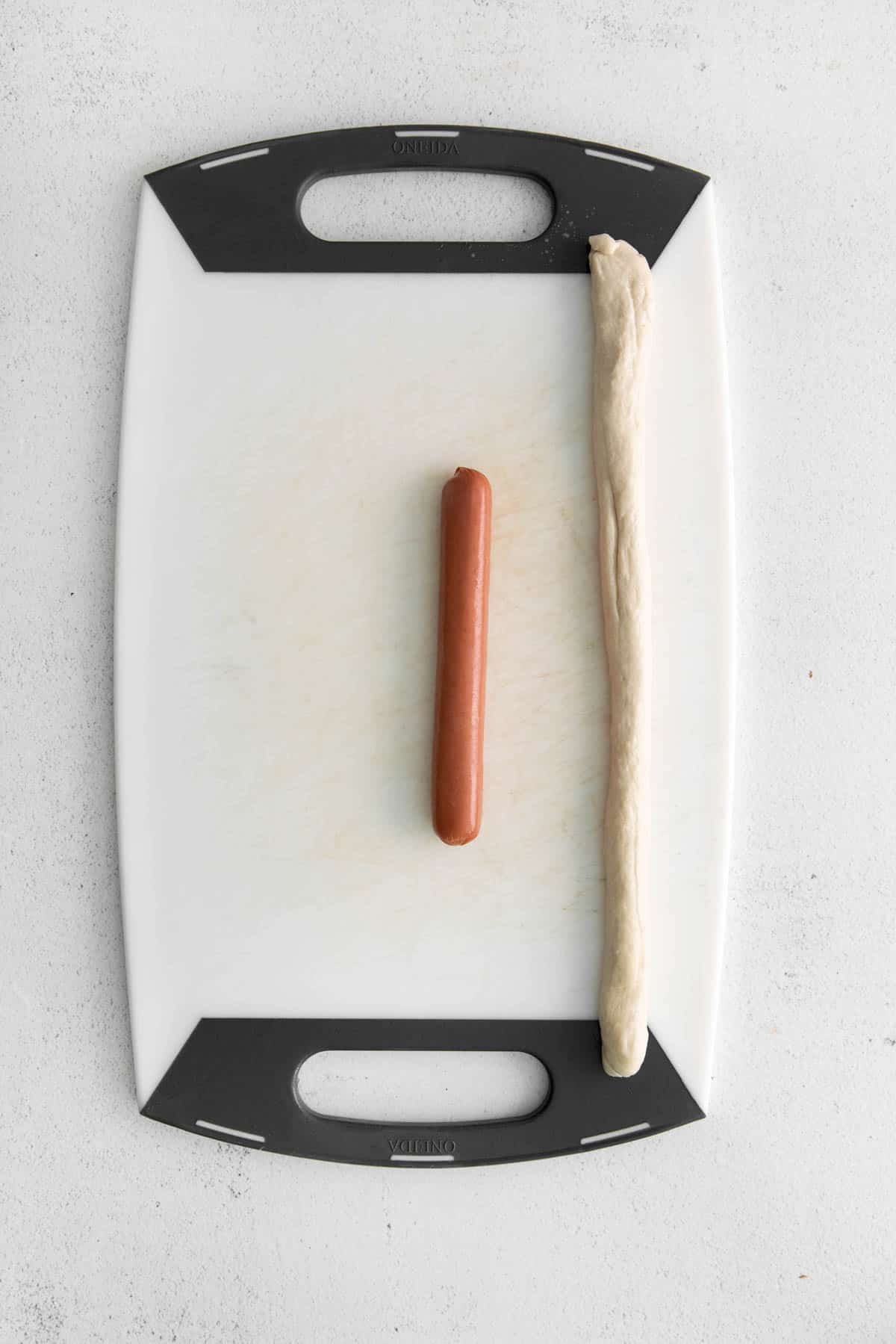 A hot dog and a piece of pretzel dough rolled into a rope on a cutting board.