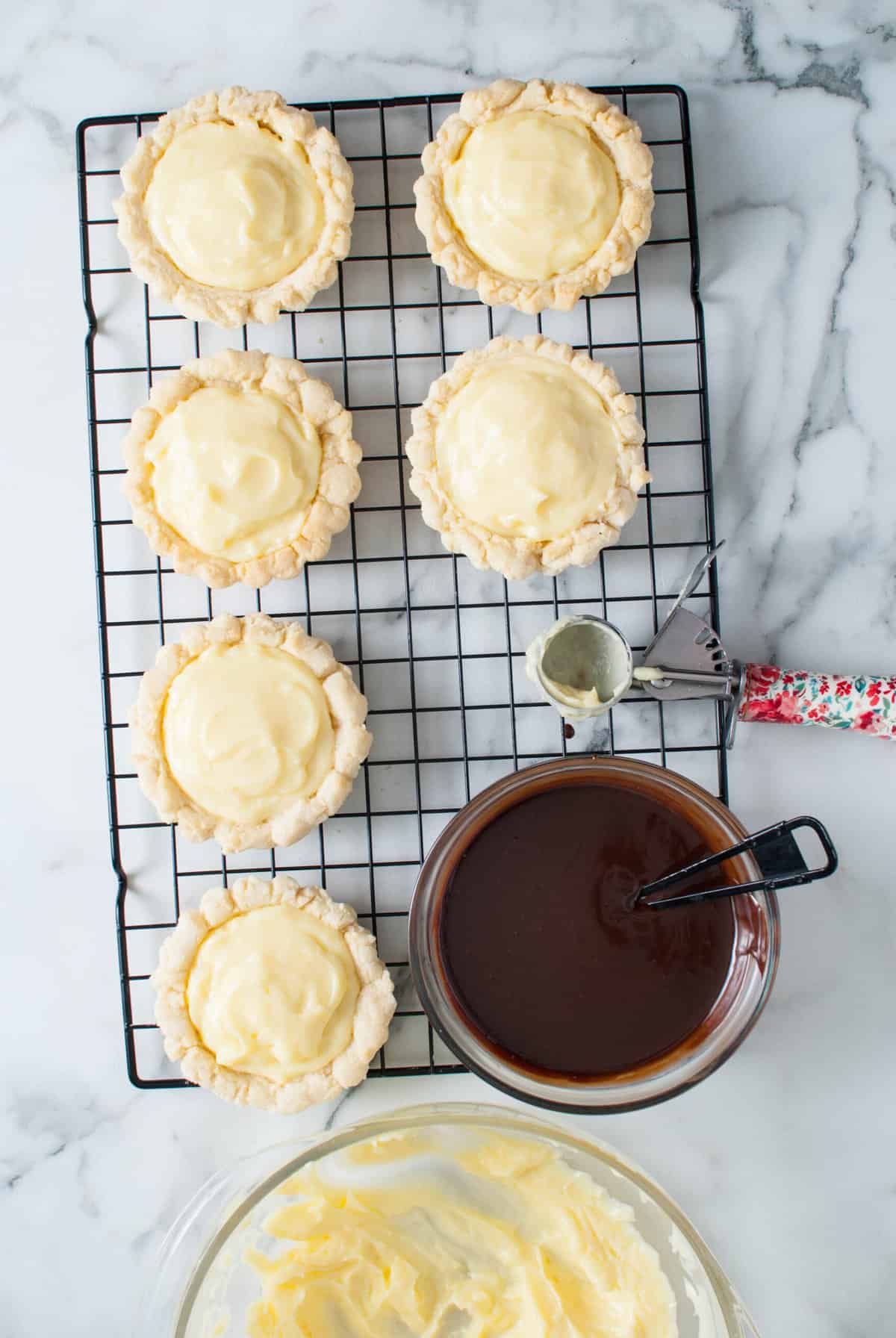 Cookie shells filled with pastry cream on a wire rack next to a bowl of chocolate glaze.