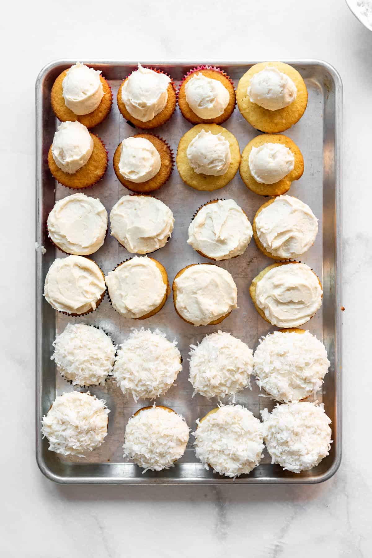 Coconut cupcakes on a baking sheet in various stages of being decorated with frosting and coconut.