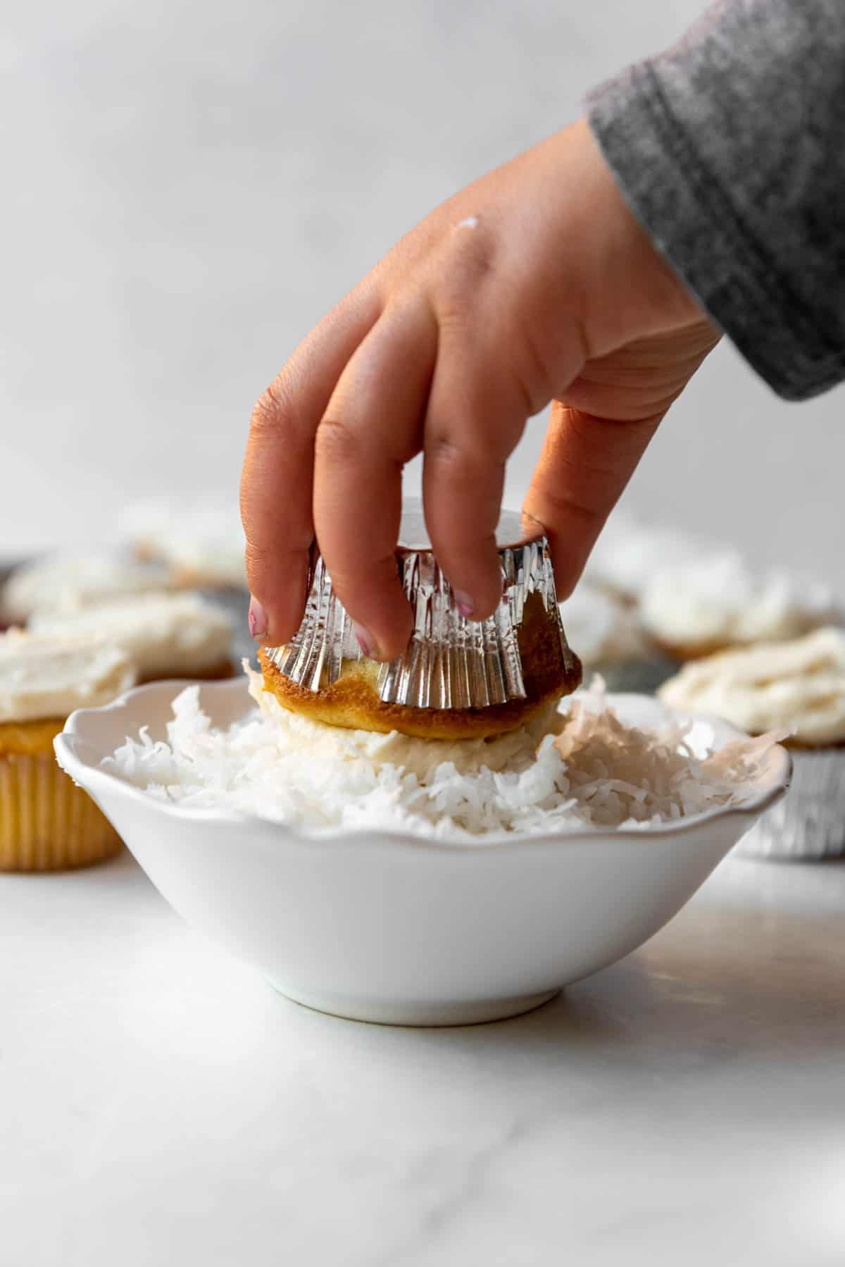 A hand dipping a frosted coconut cupcake into a bowl of shredded sweetened coconut.