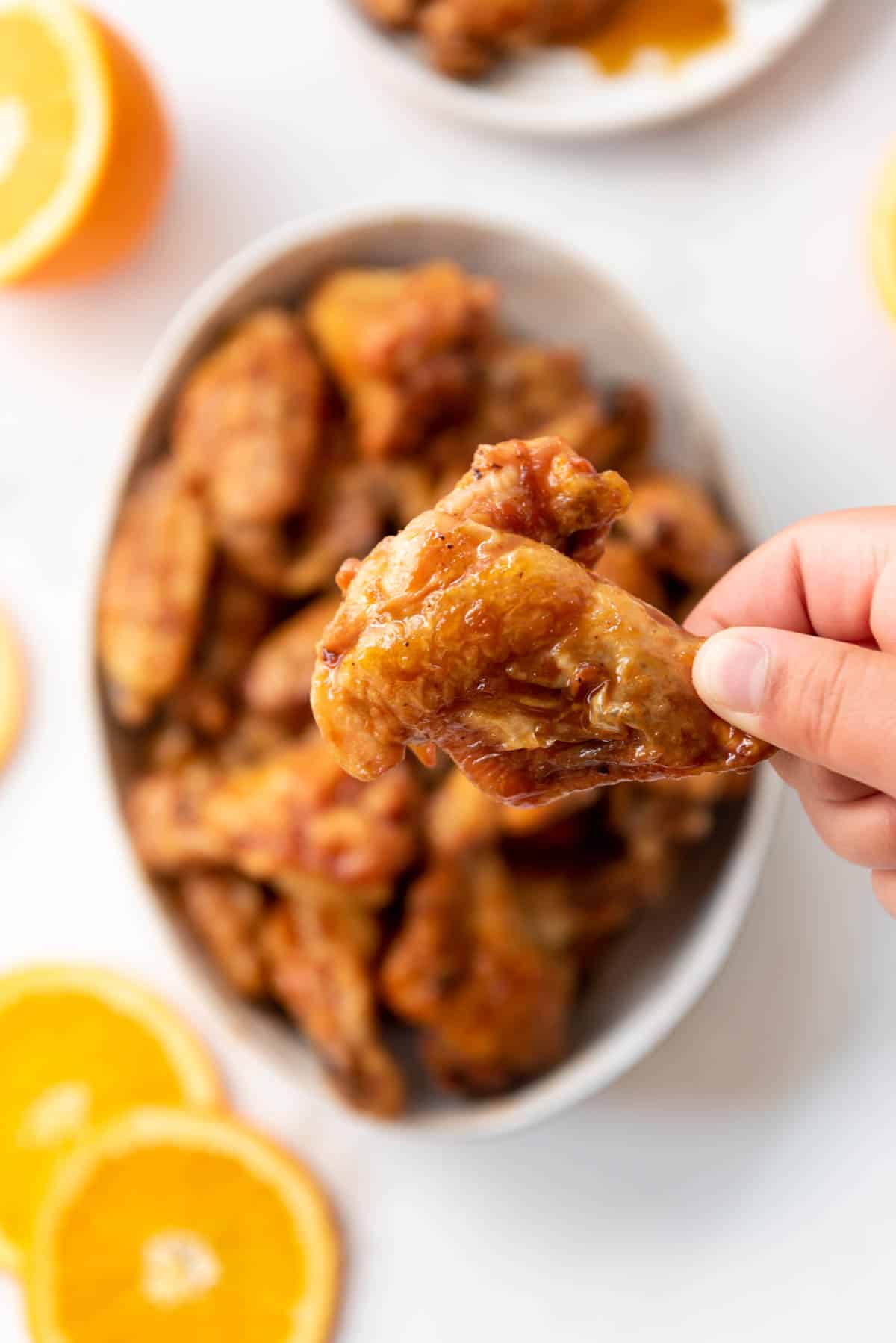 A hand holding a crispy orange chicken wing over a bowl of wings.