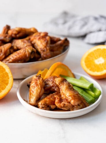 A plate of orange glazed chicken wings with celery sticks on the plate in front of a bowl of more wings.