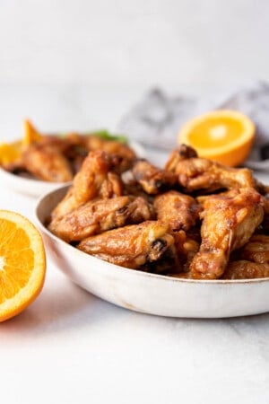 A bowl of orange glazed chicken wings next to orange halves.