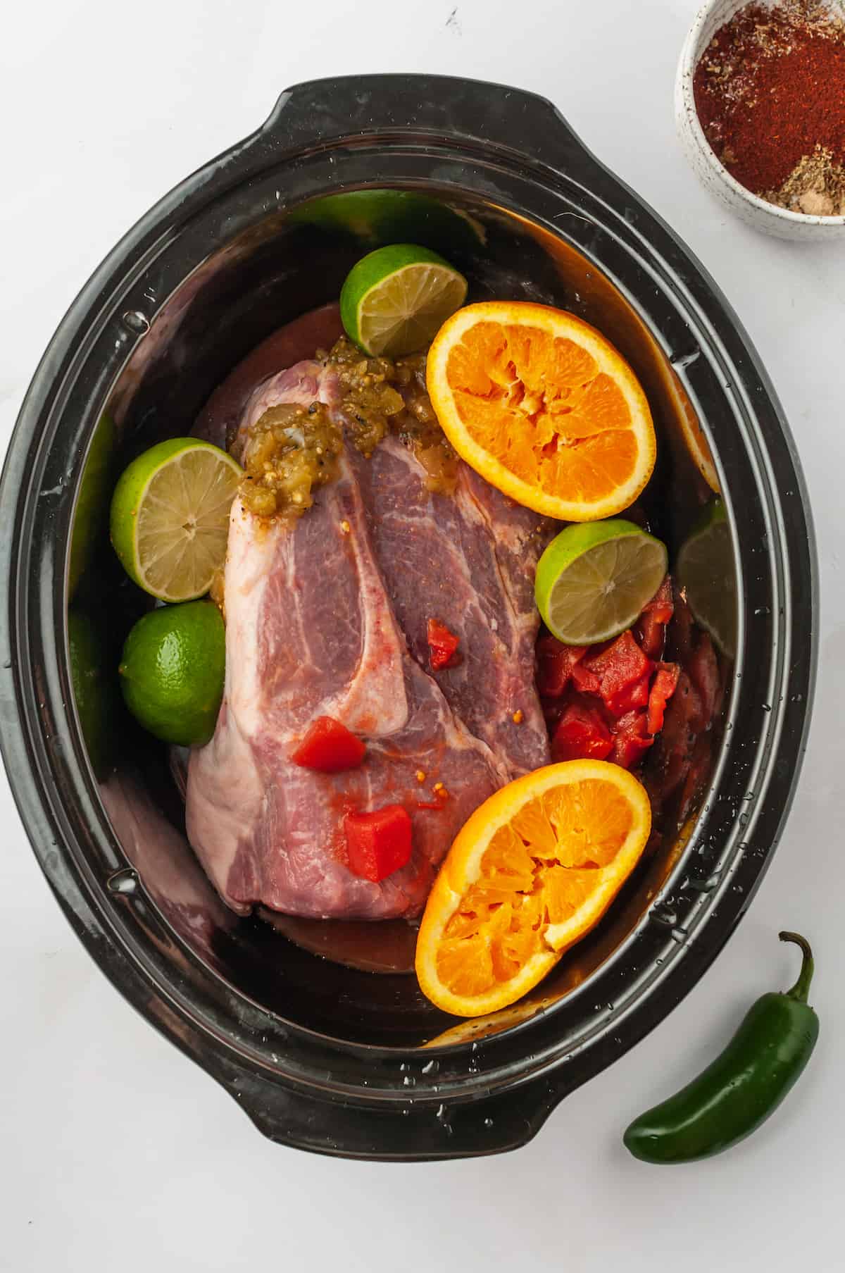 Overhead view of pork in slow cooker with tomatoes, limes, and oranges