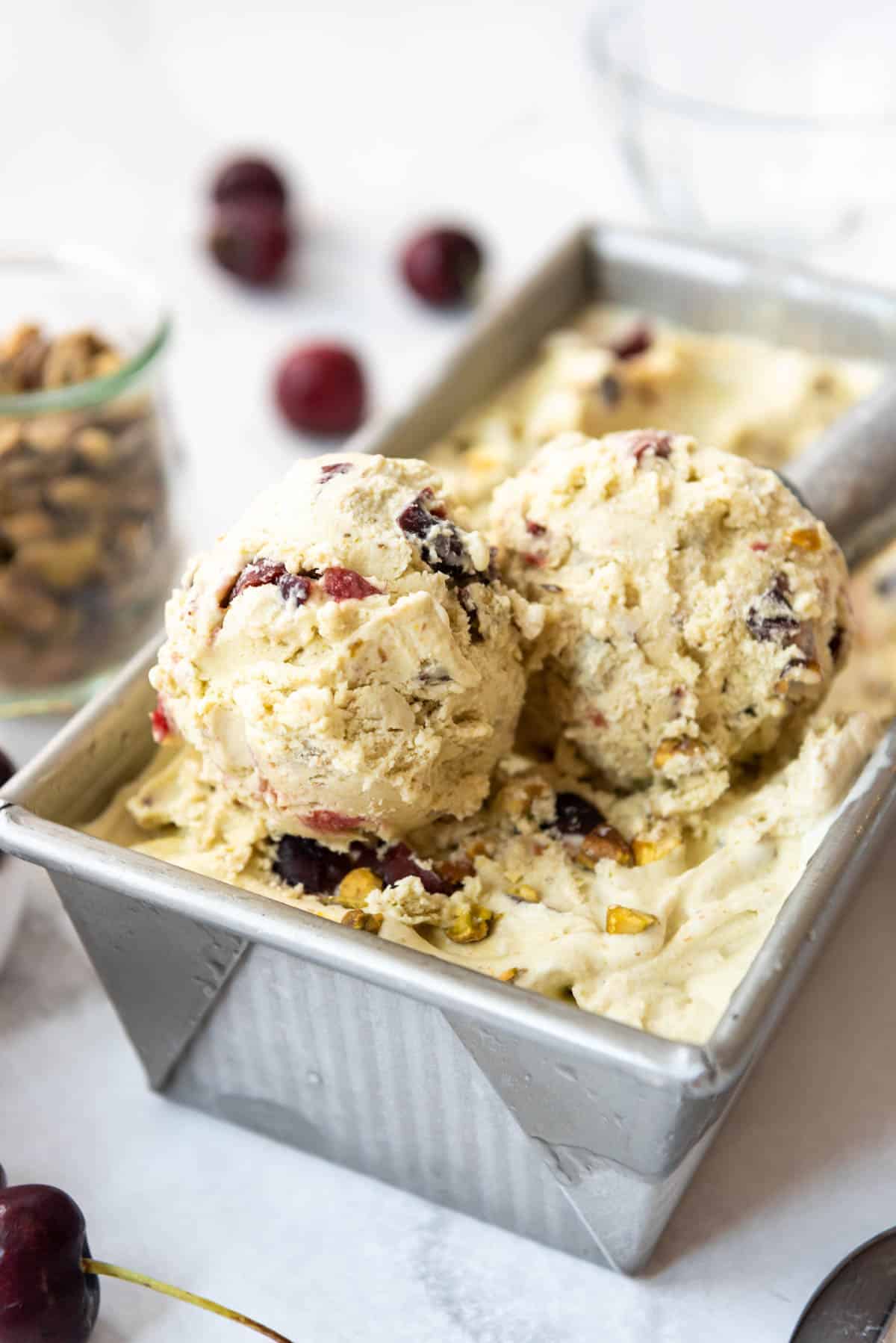 Scoops of cherry pistachio ice cream in a bread pan with cherries and pistachios nearby.