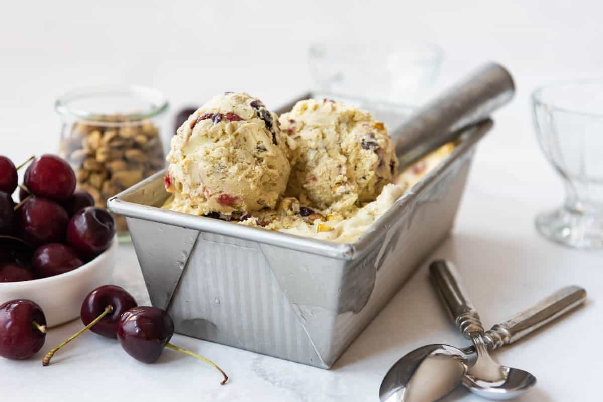 A bread pan filled with cherry pistachio ice cream with two scoops on top next to spoons and fresh sweet cherries.