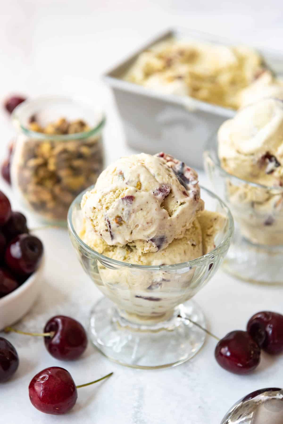 A scoop of cherry pistachio ice cream in a glass dish in front of more ice cream surrounded by pistachios and cherries.