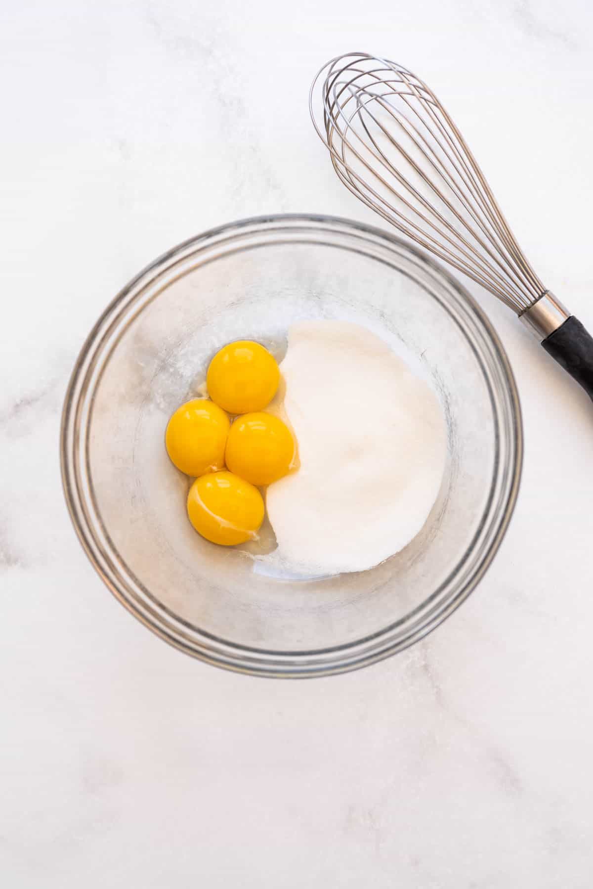 Egg yolks and granulated sugar in a glass bowl with a whisk beside it.