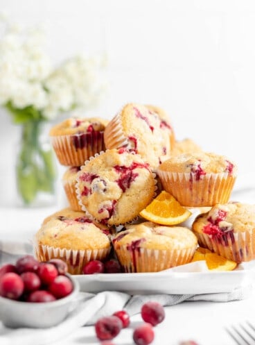 A plate of cranberry orange muffins next to a bowl of fresh cranberries in front of a vase of flowers.