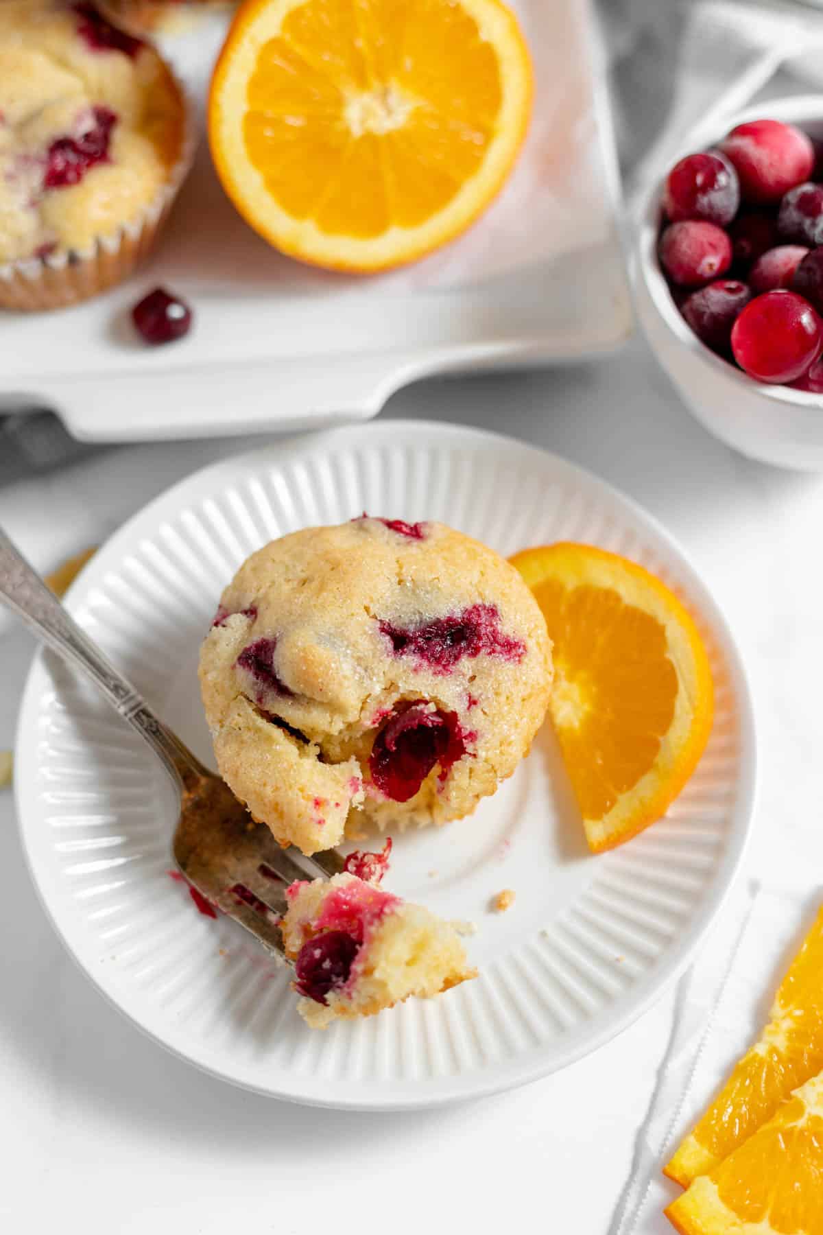 A cranberry orange muffin on a white plate with a fork with a bite of muffin on it and a slice of orange.