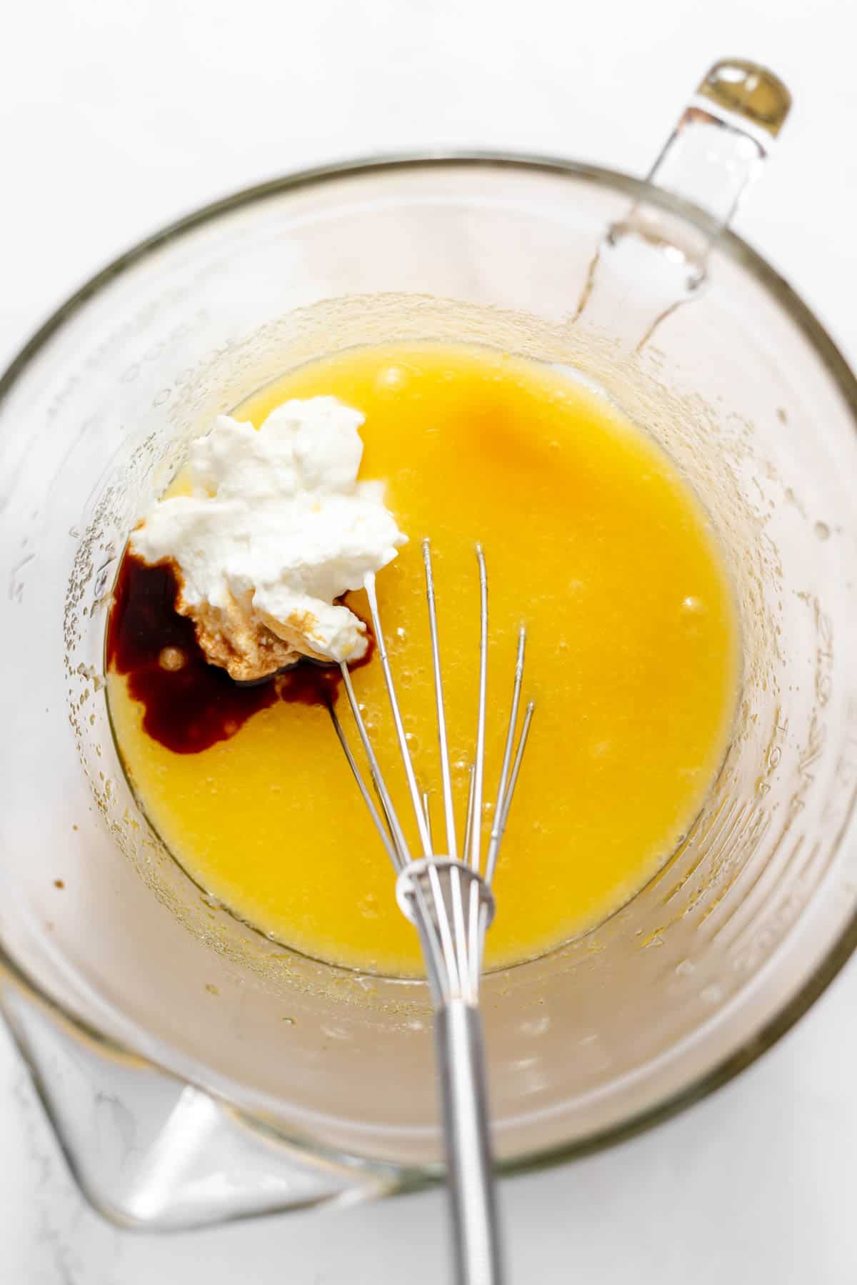 A whisk in a bowl of eggs, oil, and sugar, with vanilla and sour cream.