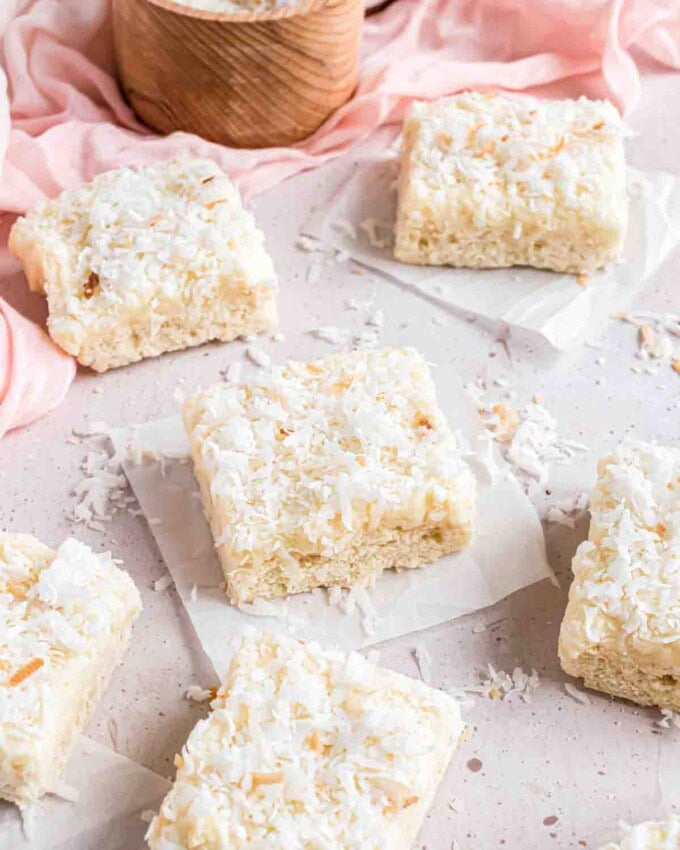 Coconut bar squares on pieces of parchment paper in front of a pink linen napkin and a wooden bowl with coconut.