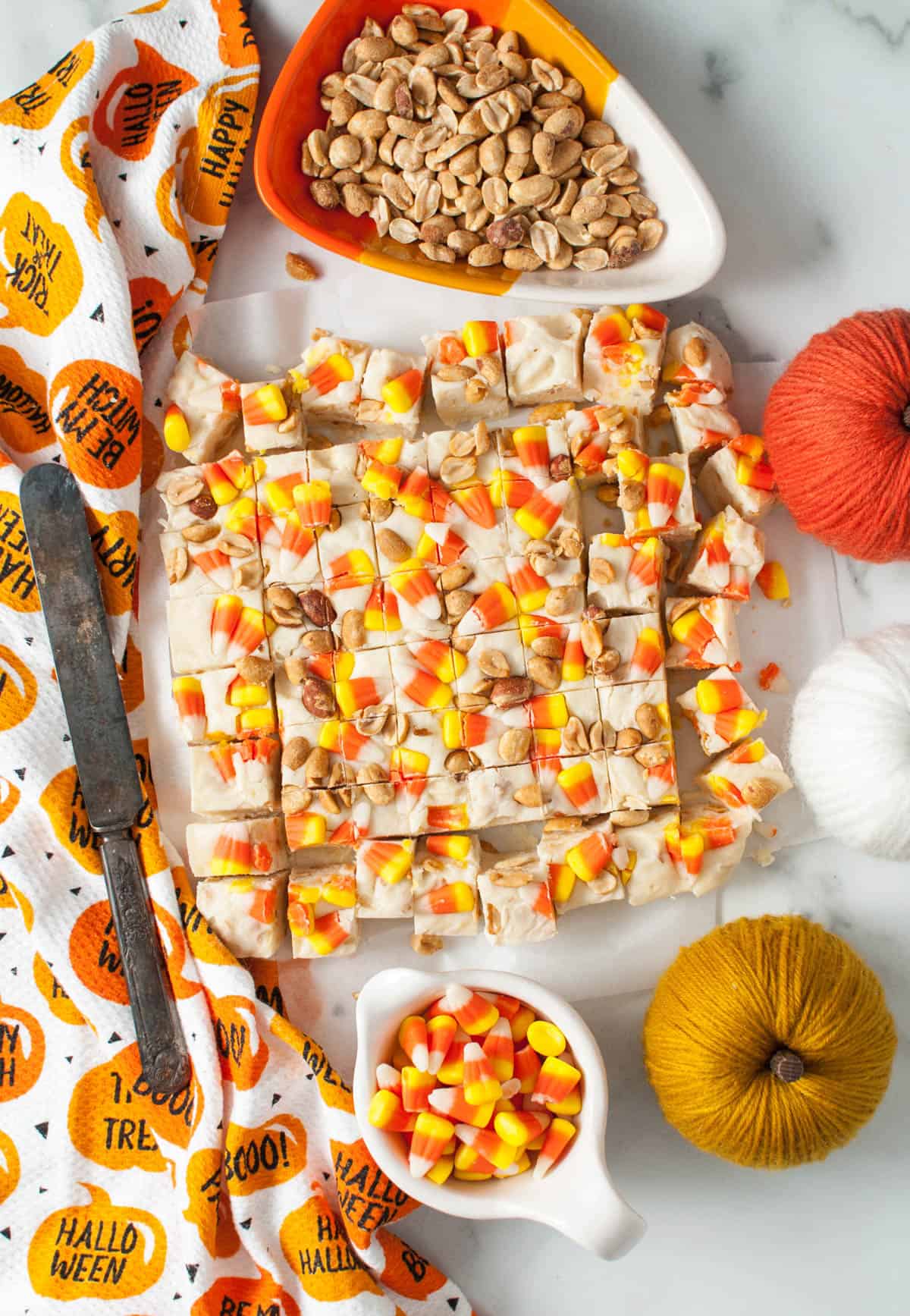 An overhead image of a batch of candy corn fudge cut into small squares beside bowls of roasted peanuts and candy corn.