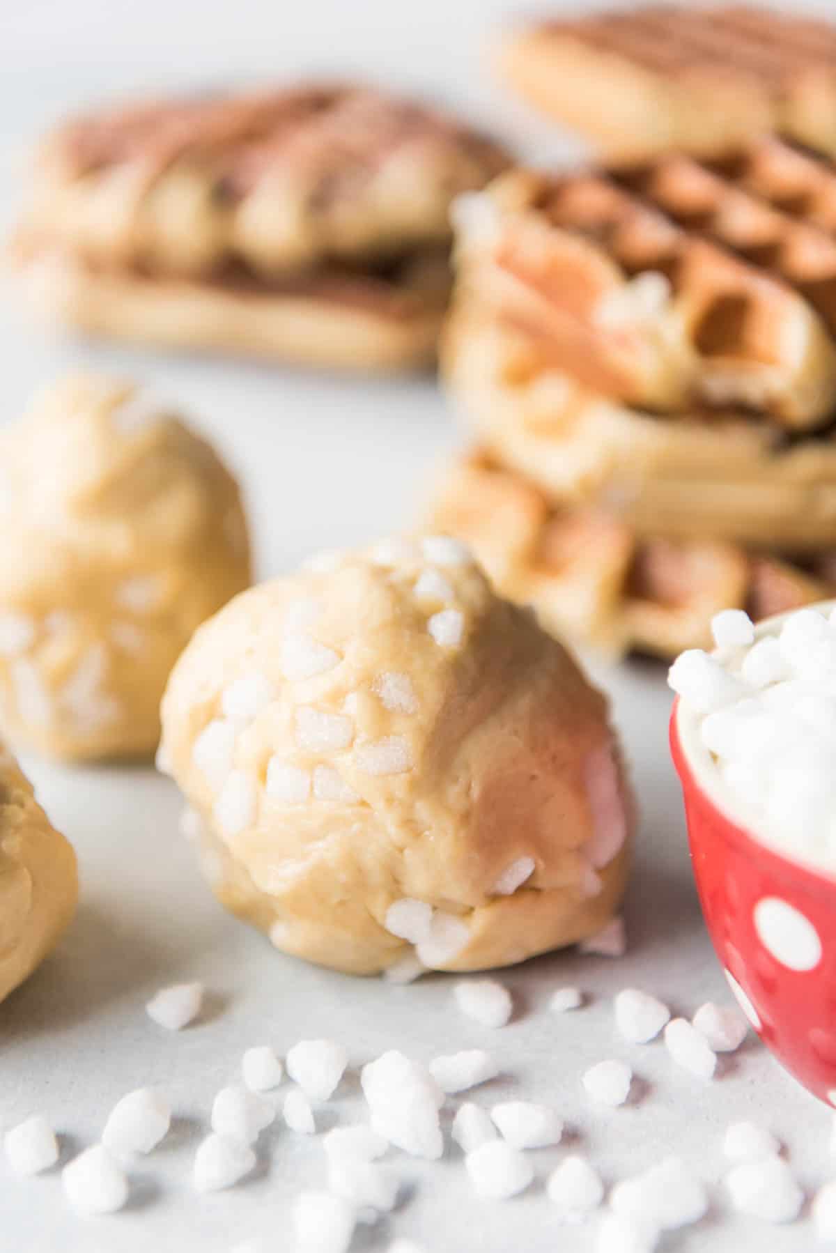 Balls of liege waffle dough with sugar pearls next to stacks of waffles.