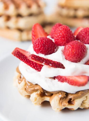A waffle with sugar pearls topped with cream and fresh berries with more waffles in the background.