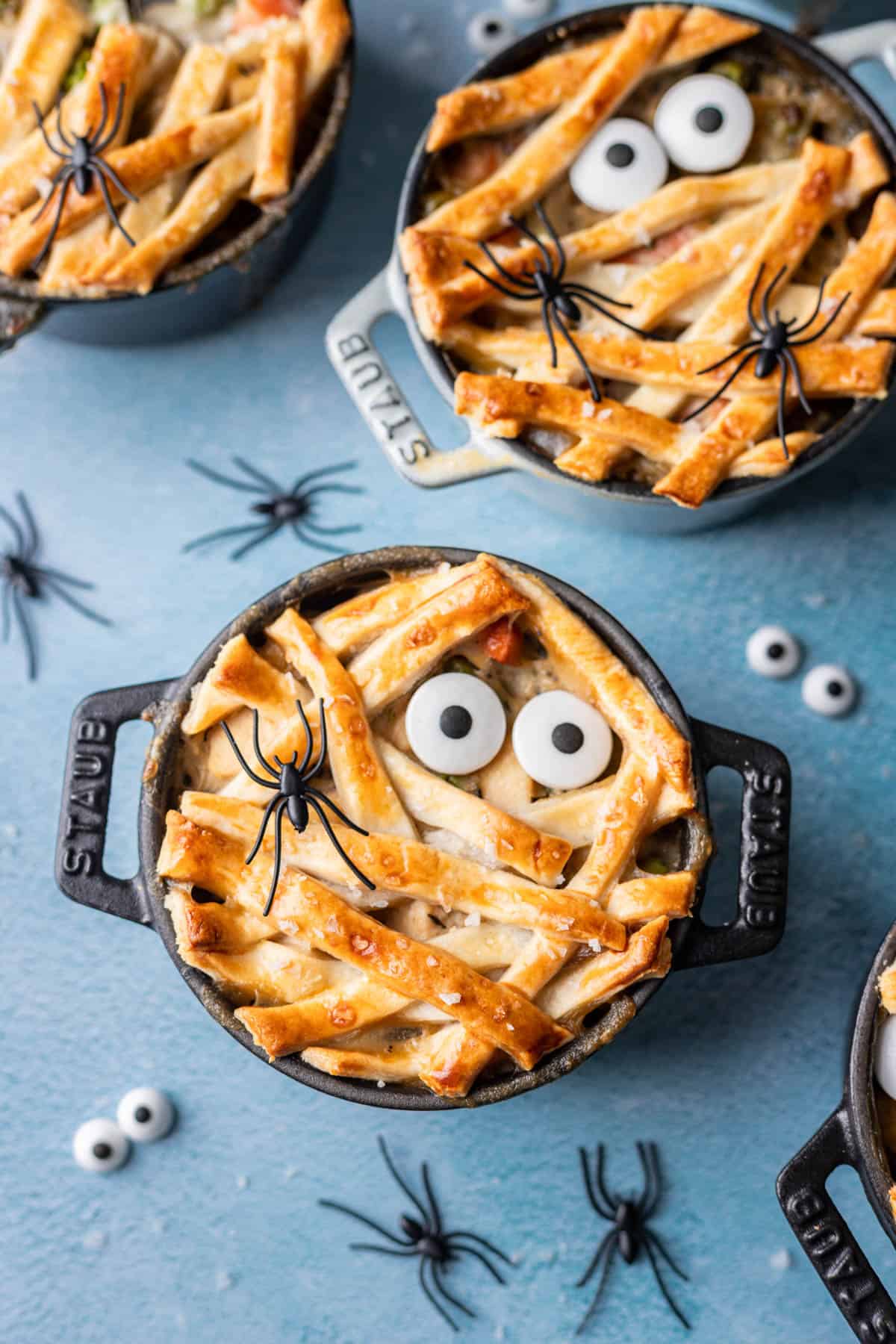 Mini chicken pot pies decorated like mummies for Halloween with plastic spiders and candy eyeballs on a blue background.