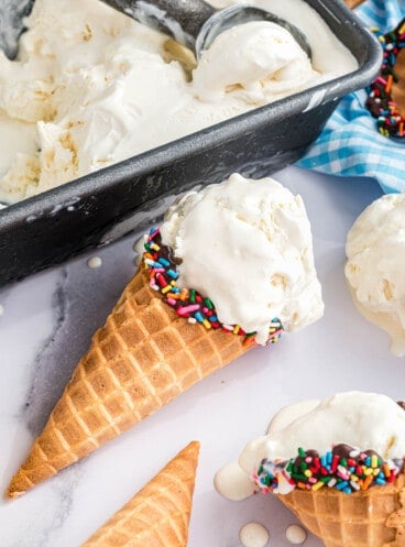 A scoop of no-churn vanilla ice cream in a waffle cone laying on a white surface next to more ice cream cones and a pan of homemade vanilla ice cream.