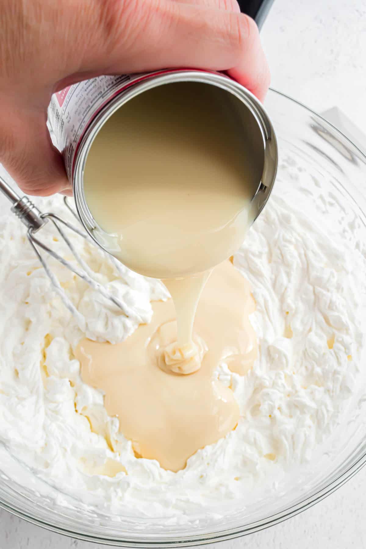 A hand pouring sweetened condensed milk from the can into a bowl of whipped cream.