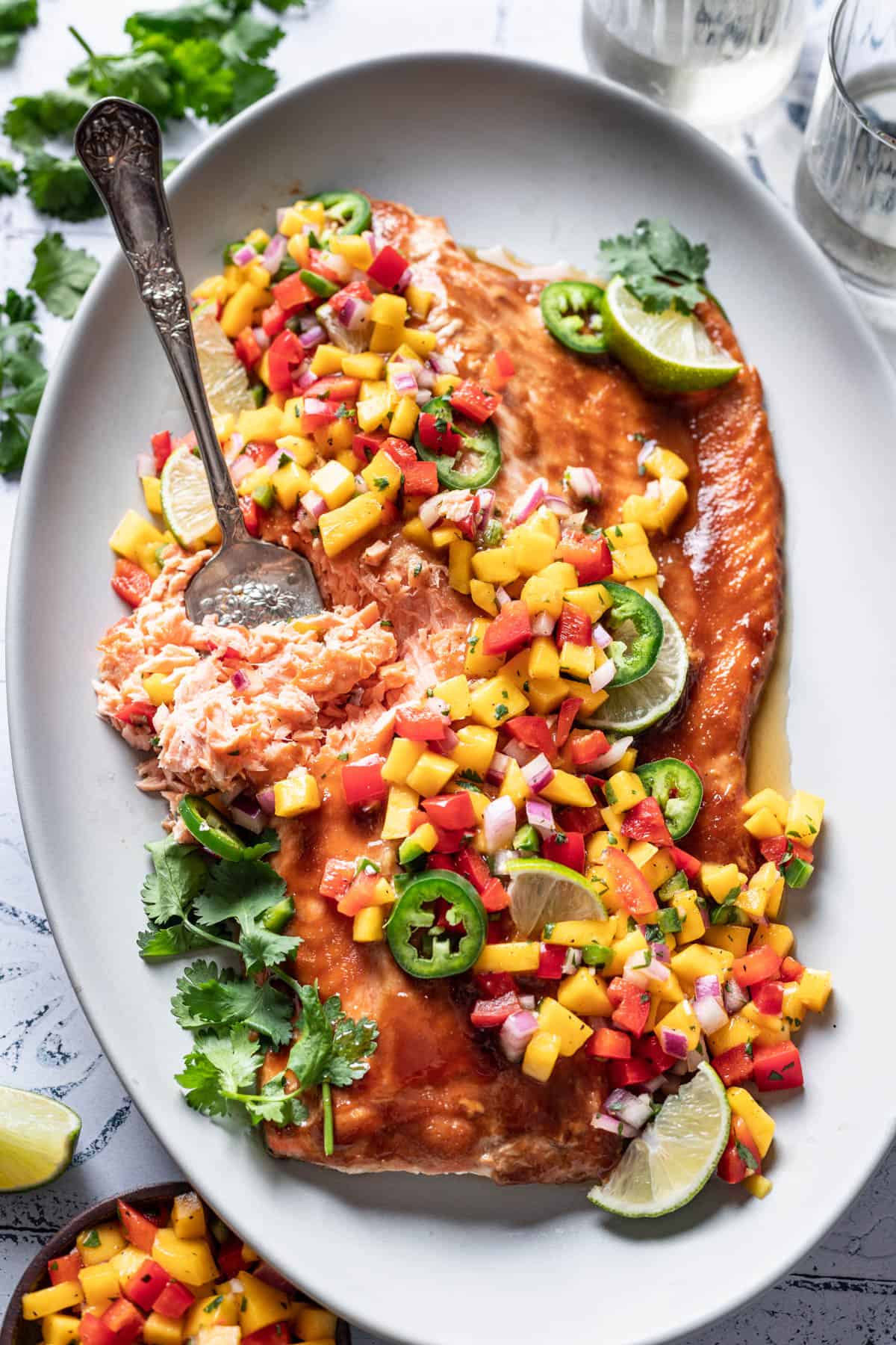 A large baked salmon fillet with mango salsa and a serving fork flaking the fish on a white serving platter.