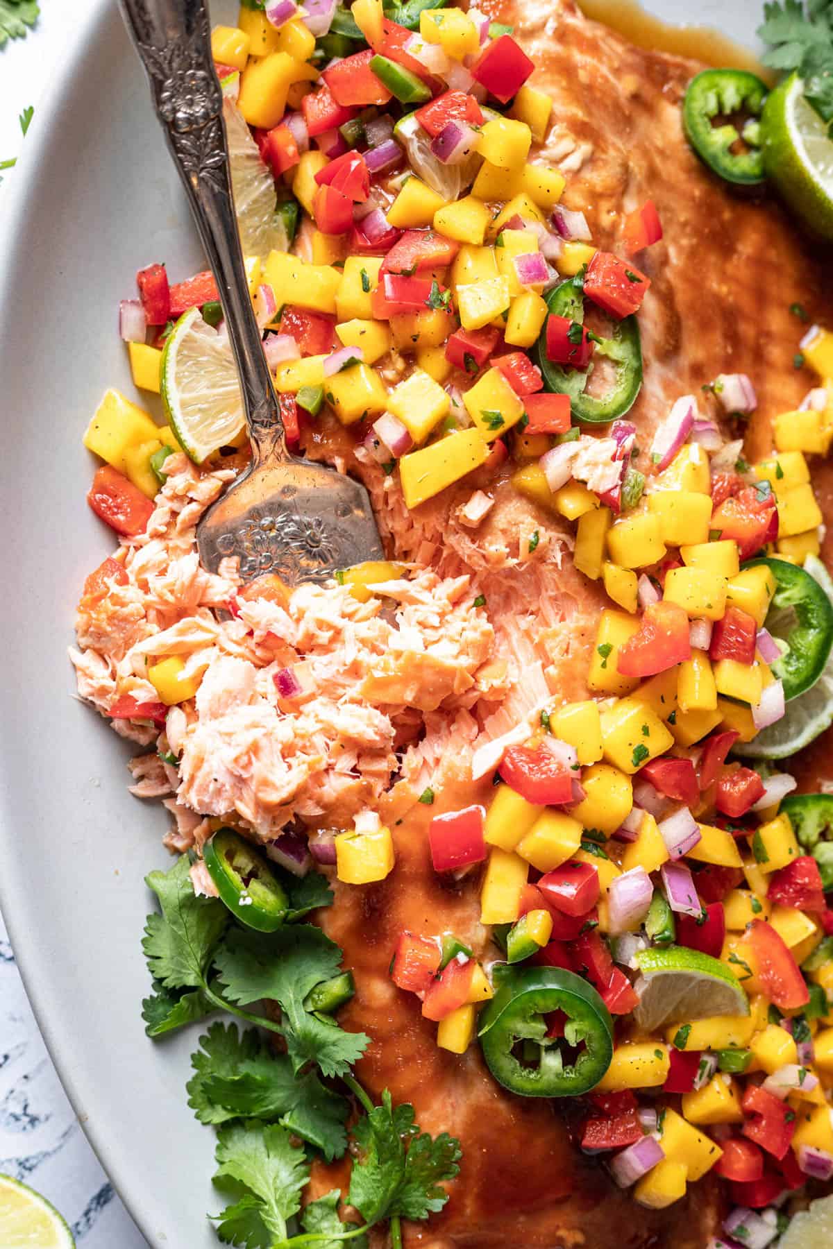 Close up of a large fillet of salmon with mango salsa on top, served on a white plate with a fork in it flaking off some pieces of salmon.