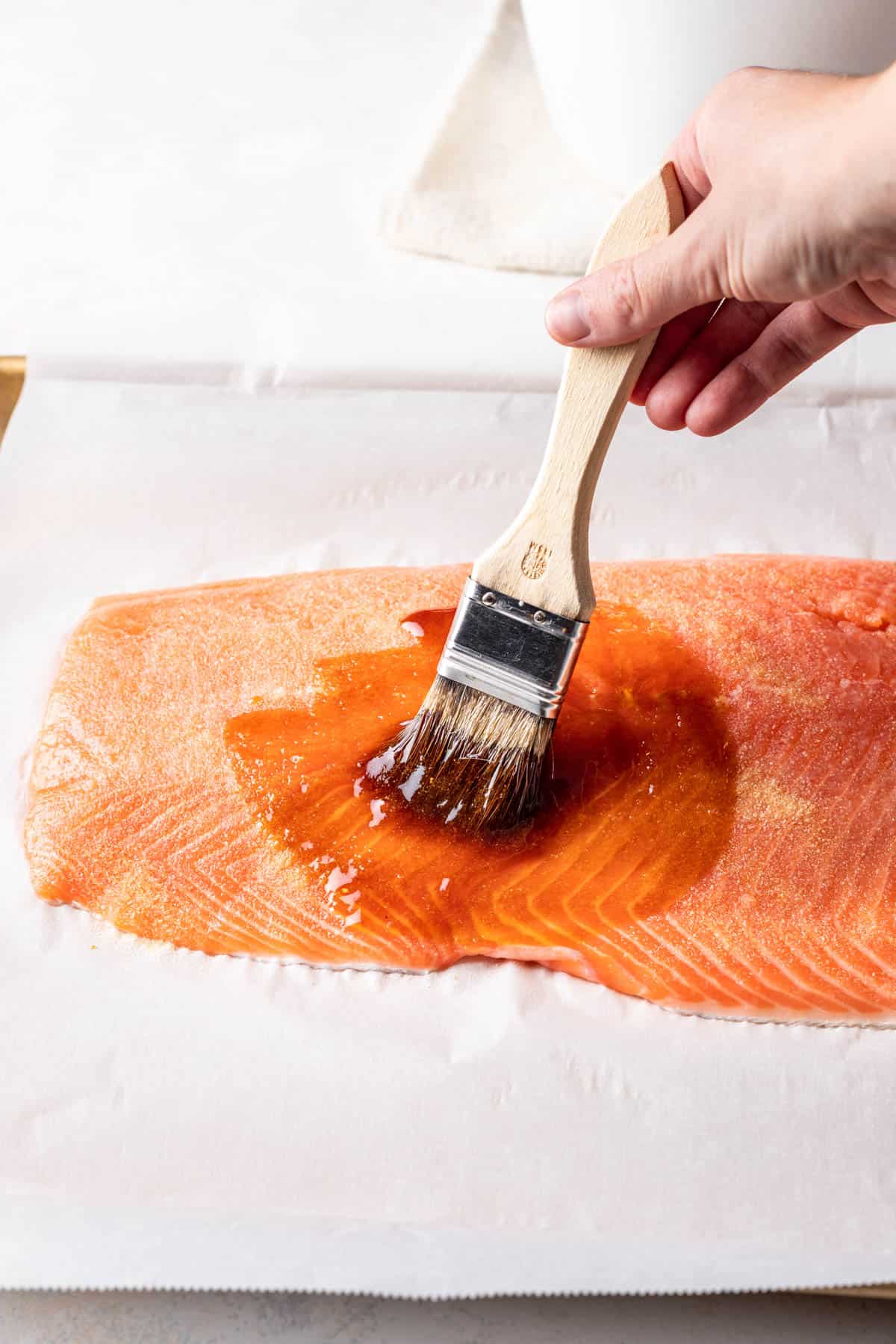 A hand brushing maple soy glaze on a piece of salmon with a pastry brush.