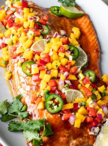 A large salmon fillet served family style on a white plate with fresh mango salsa on top.