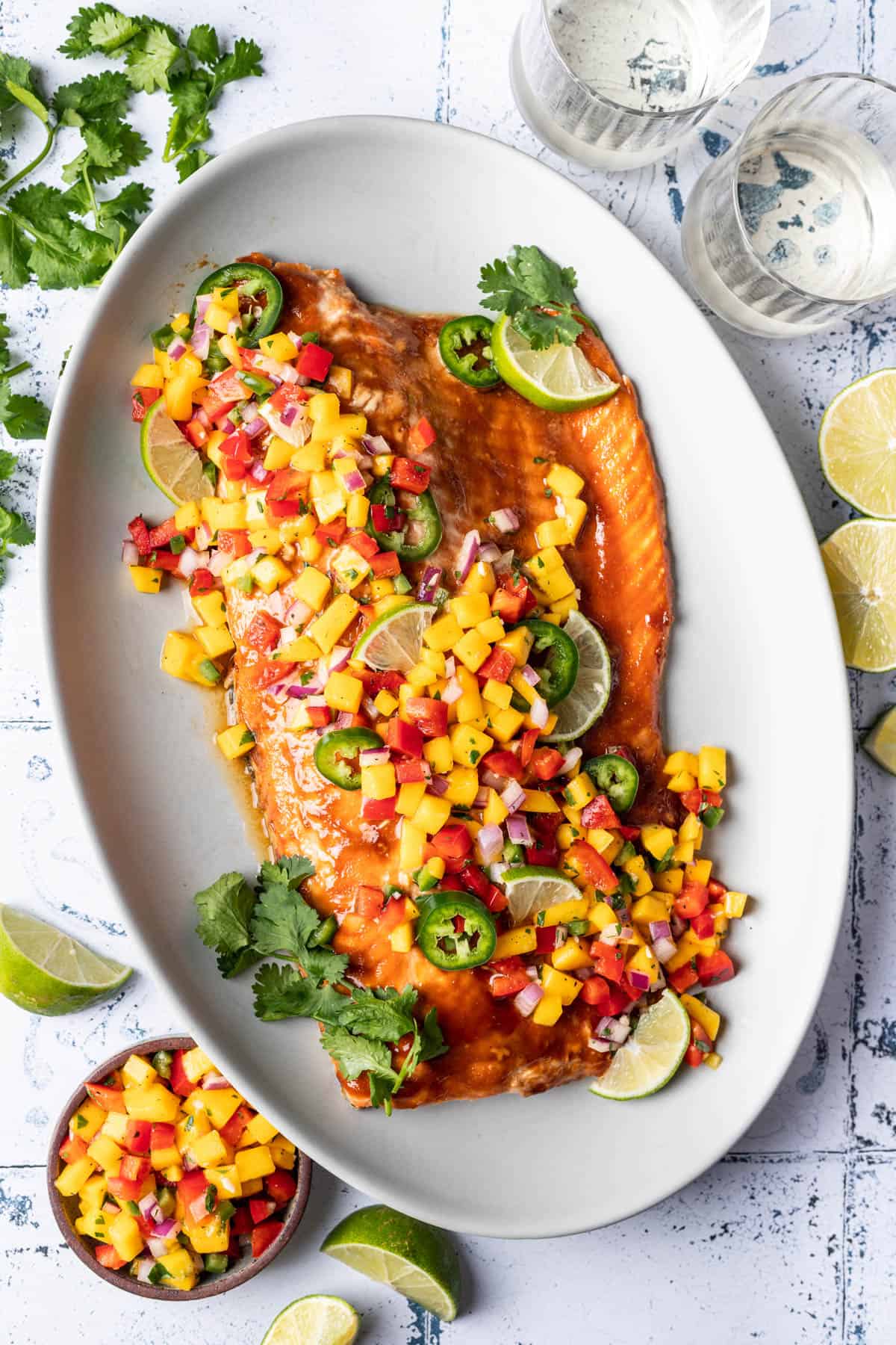 A large salmon fillet served family style on a white plate with fresh mango salsa on top.