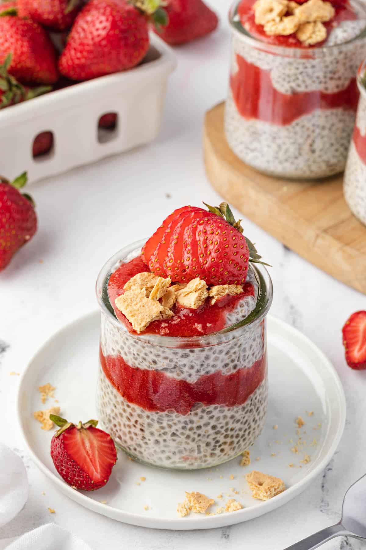 An angled image of a sliced strawberry and broken up graham crackers topping a jar of chia seed pudding.