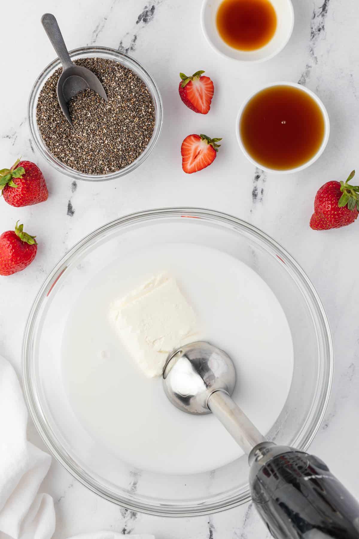 An immersion blender in a bowl of coconut milk with cream cheese next to strawberries and a bowl of chia seeds.