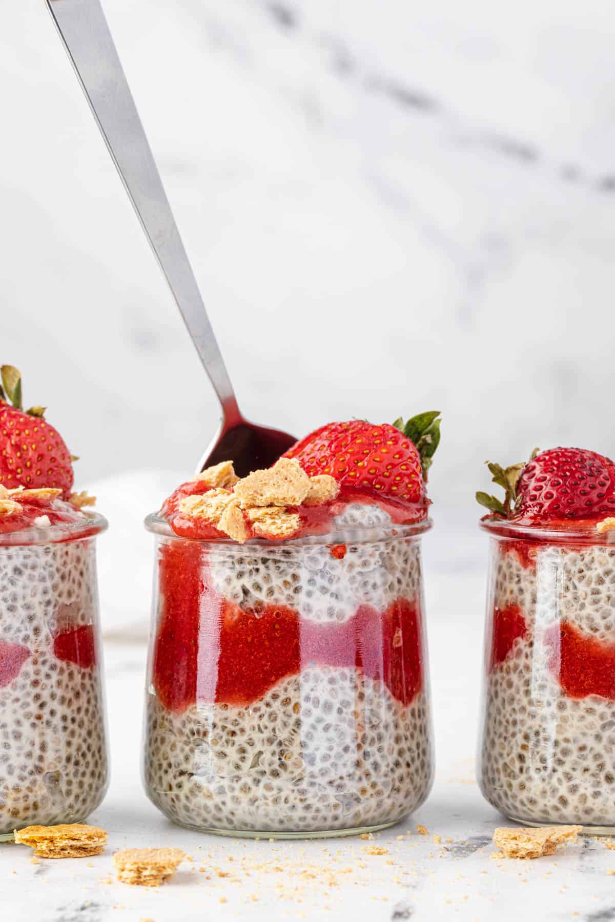 A spoon sticking out of a jar of strawberry cheesecake chia pudding next to more jars of pudding.