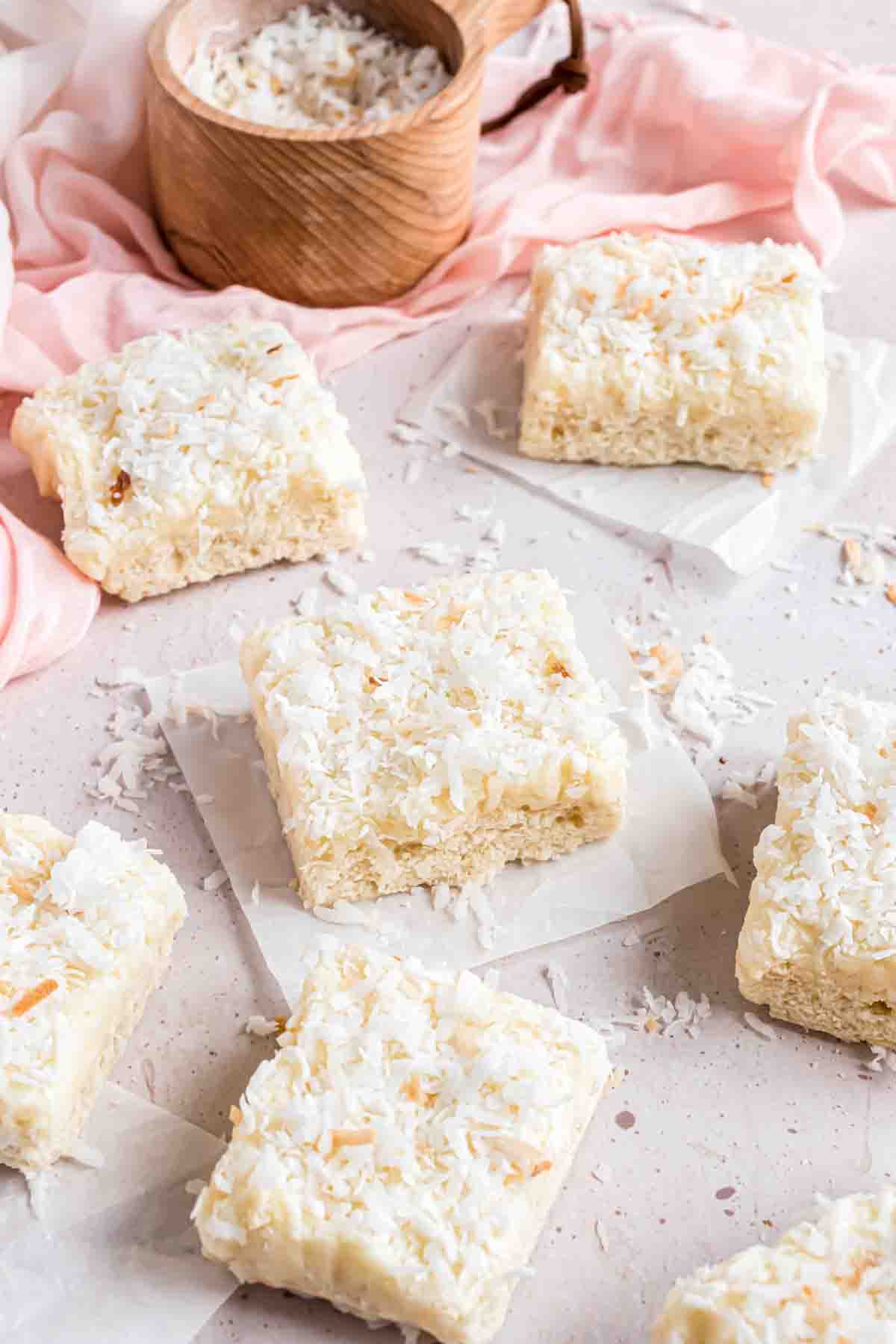 Coconut bar squares on pieces of parchment paper in front of a pink linen napkin and a wooden bowl with coconut.