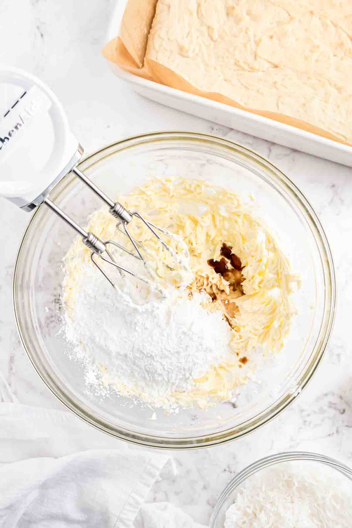 Adding powdered sugar and vanilla extract to creamed butter in a glass bowl to make frosting.