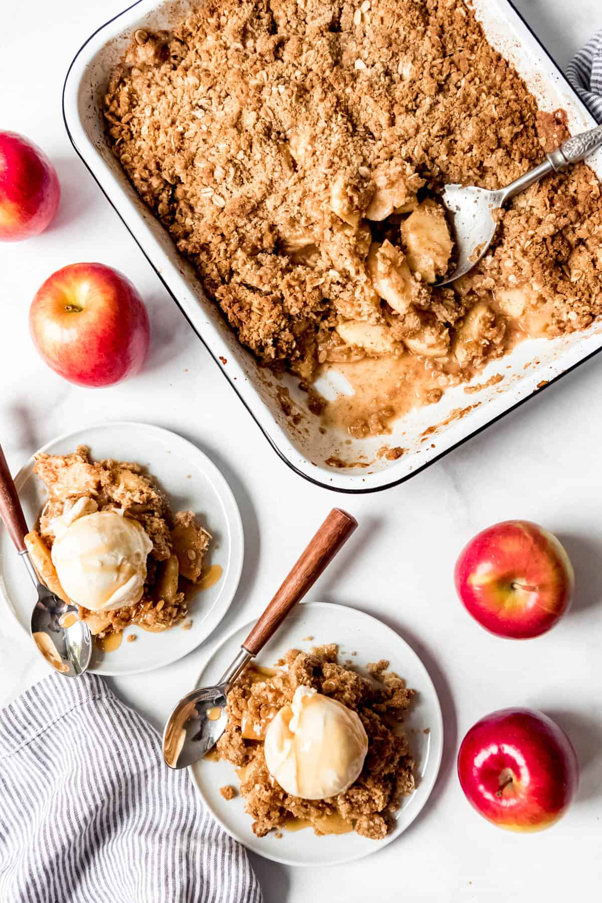 Scoops of apple crisp with vanilla ice cream on plates next to fresh apples and the pan full of the remaining apple crisp.