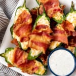 Homemade jalapeno poppers wrapped in bacon on a plate with ranch dressing.