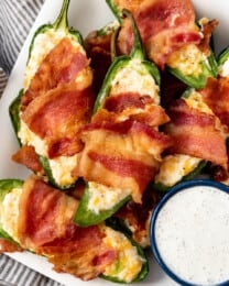 Homemade jalapeno poppers wrapped in bacon on a plate with ranch dressing.