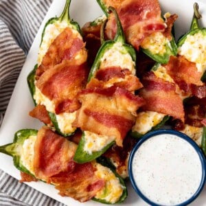 Homemade jalapeno poppers wrapped in bacon on a plate with ranch dressing.