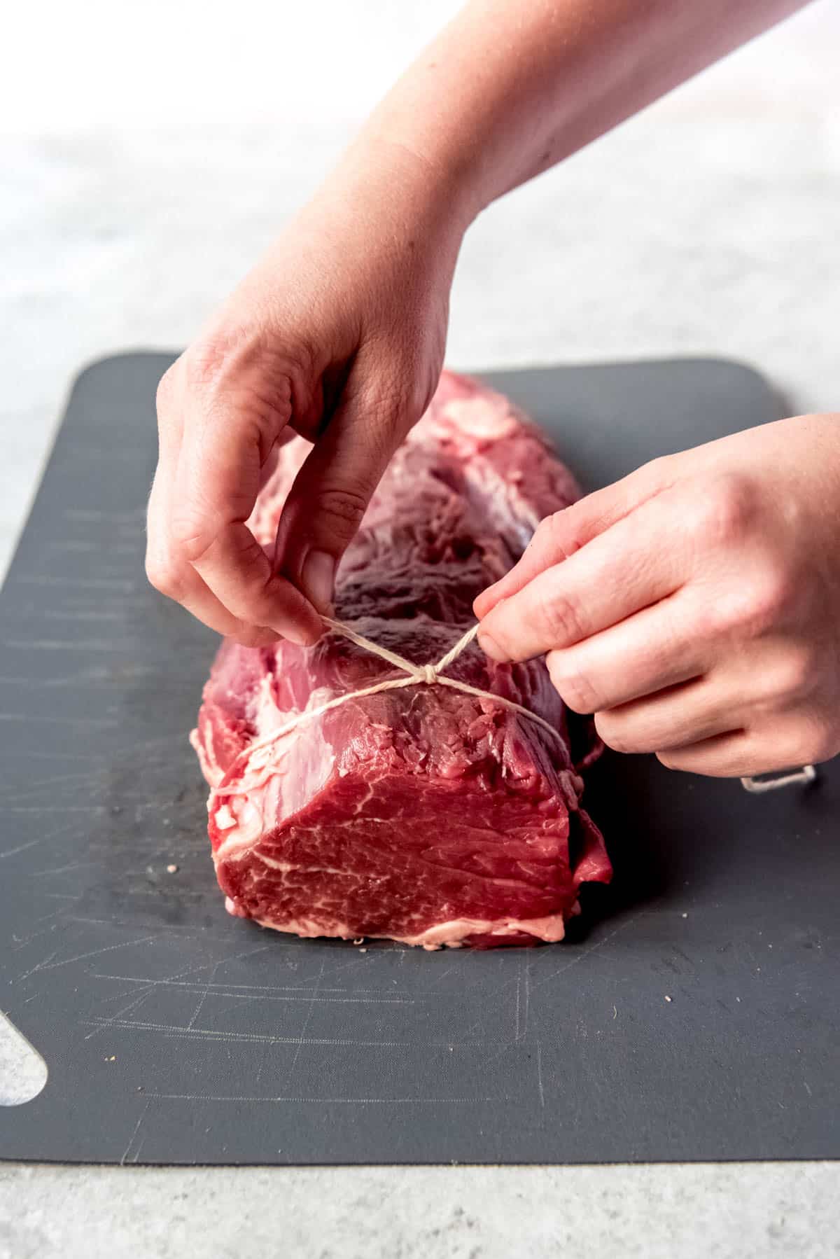 Top view of hands tying a knot in a kitchen string around a beef tenderloin roast.