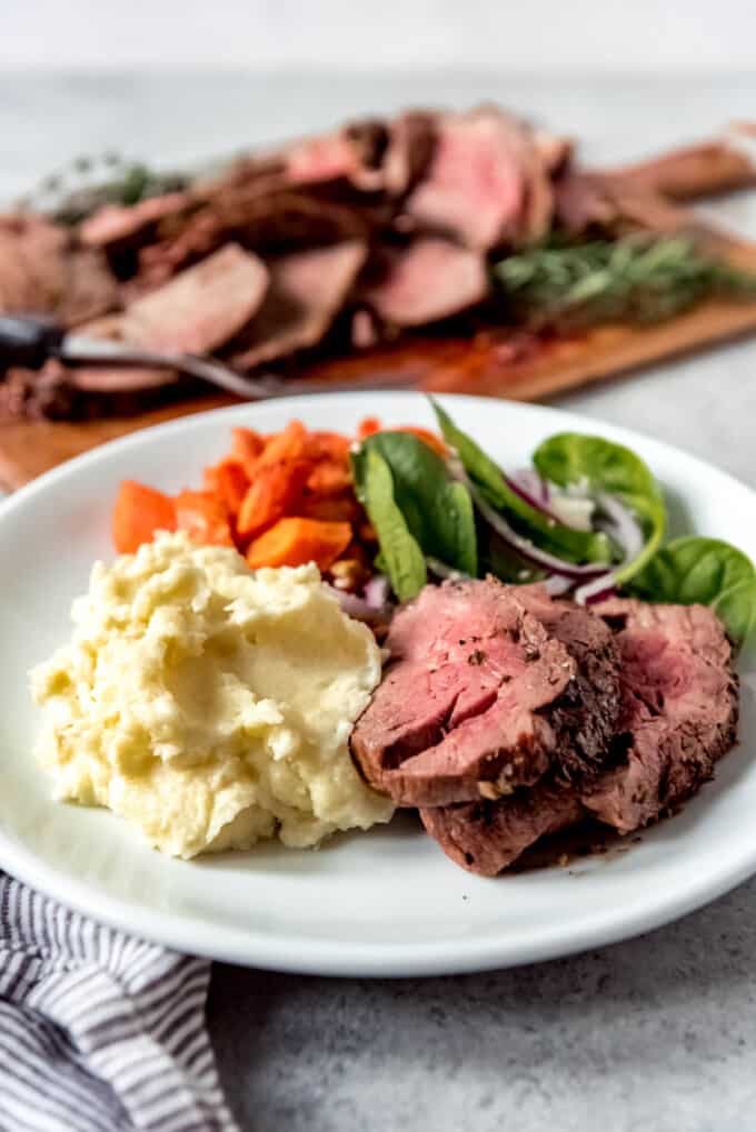 Close up of a plate with garlic herb butter roasted beef tenderloin, creamy mashed potatoes, salad, and roasted carrots.
