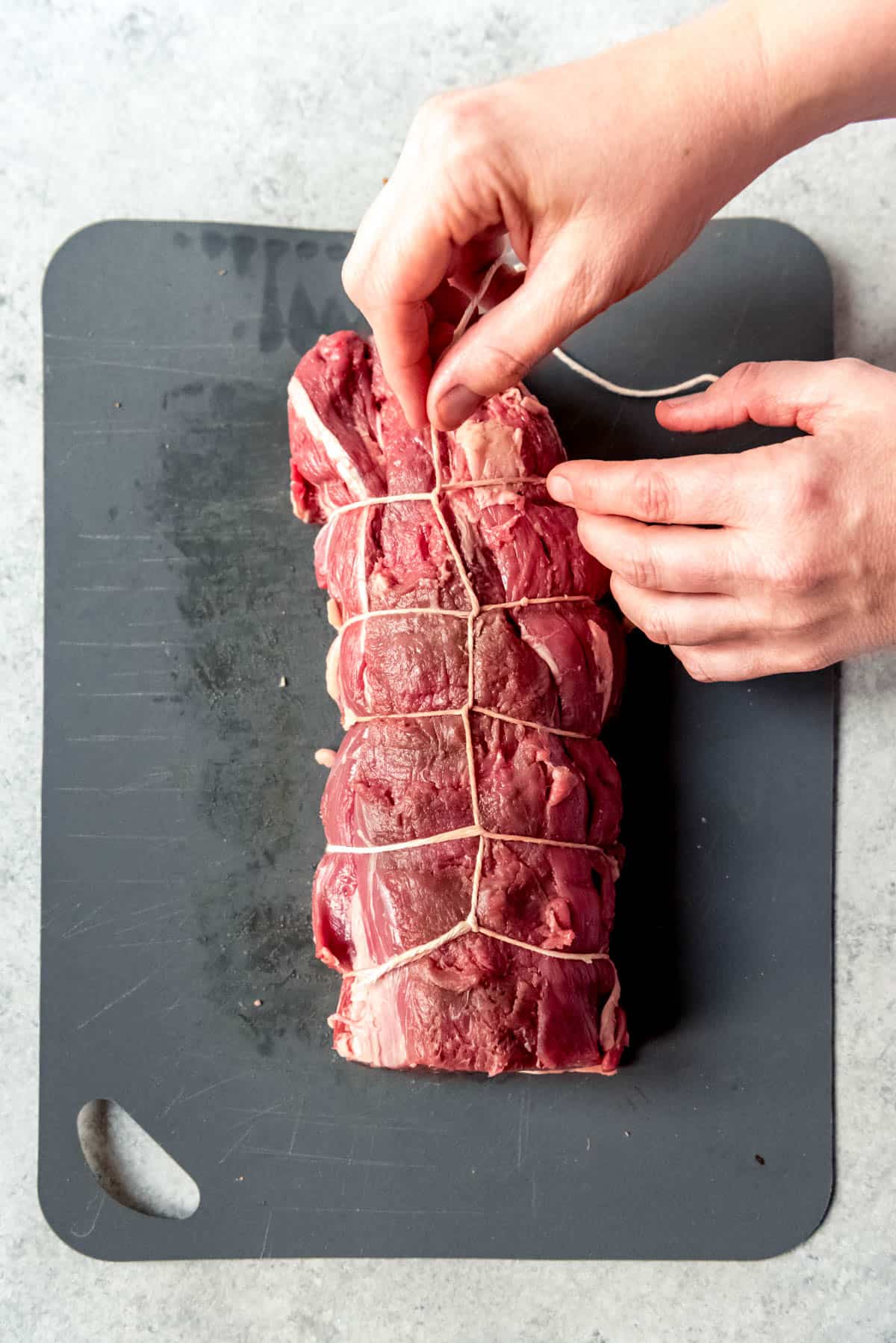 Top view of a raw beef tenderloin being wrapped in string, on a chopping board. 