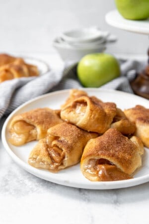 A stack of caramel apple pie crescent rolls on a white plate with a green apple in the background.