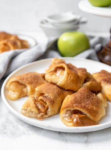 A stack of caramel apple pie crescent rolls on a white plate with a green apple in the background.