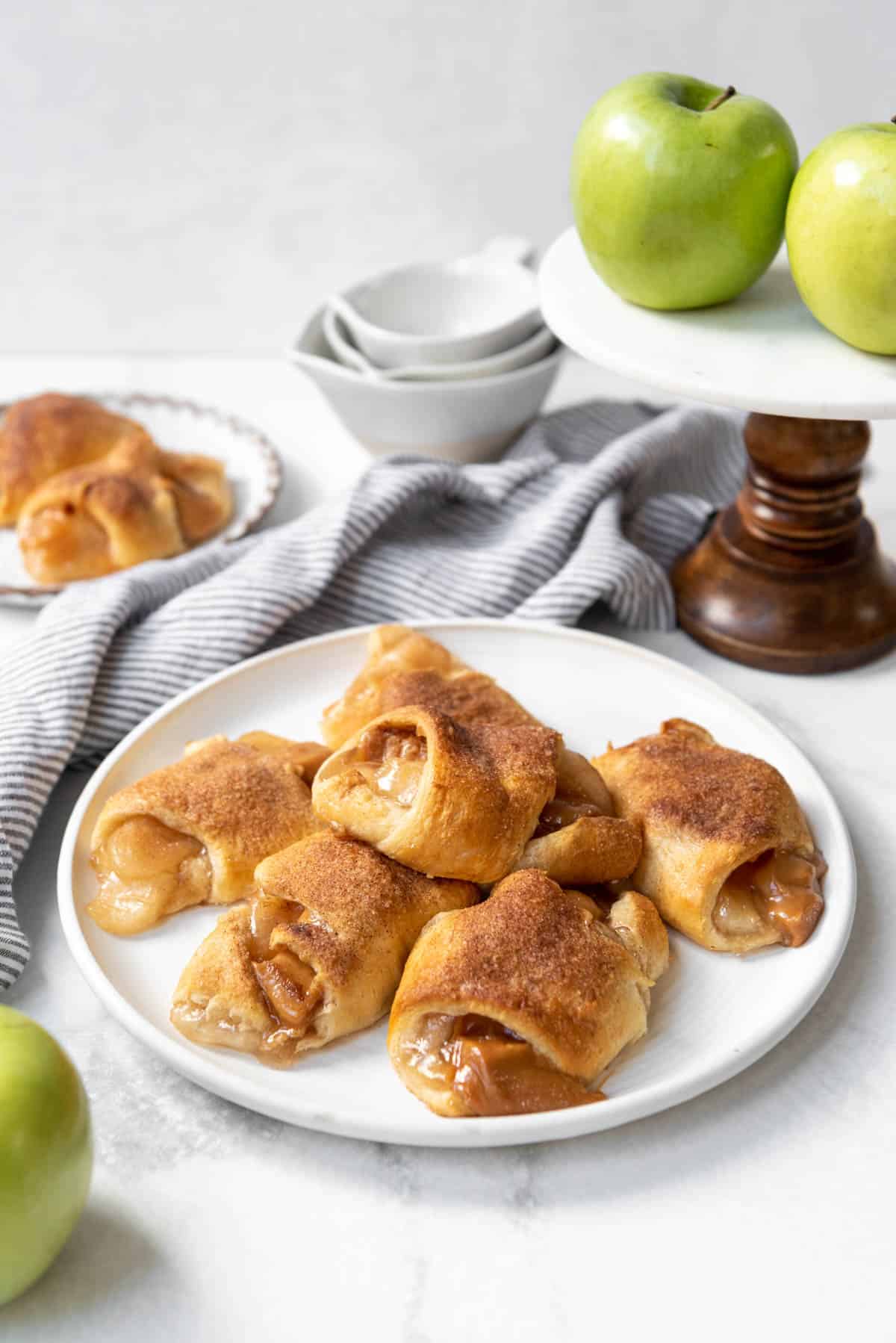 A plate of caramel apple pie crescent rolls in front of a cake stand with Granny Smith apples.