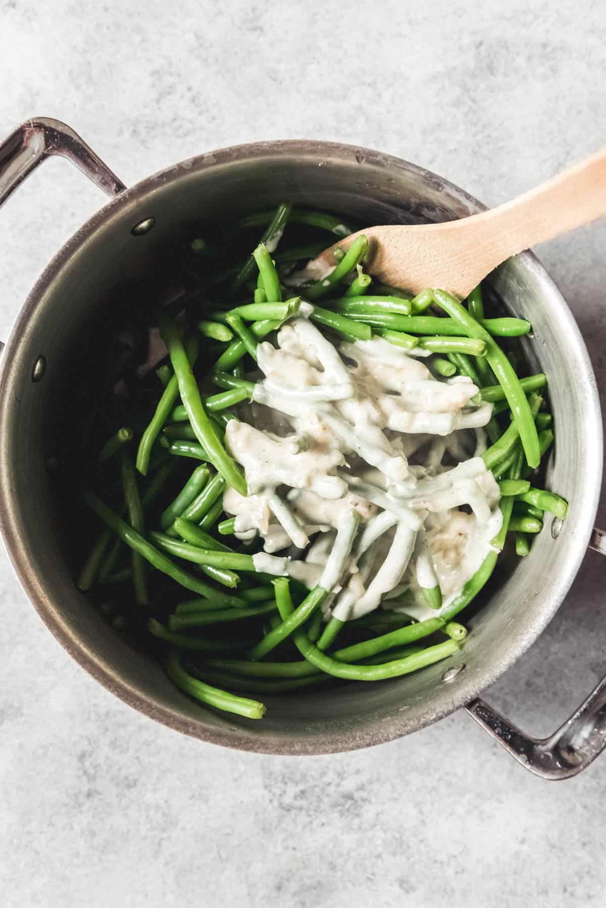 Adding a creamy sauce to cooked green beans in a large pot with a wooden spoon.