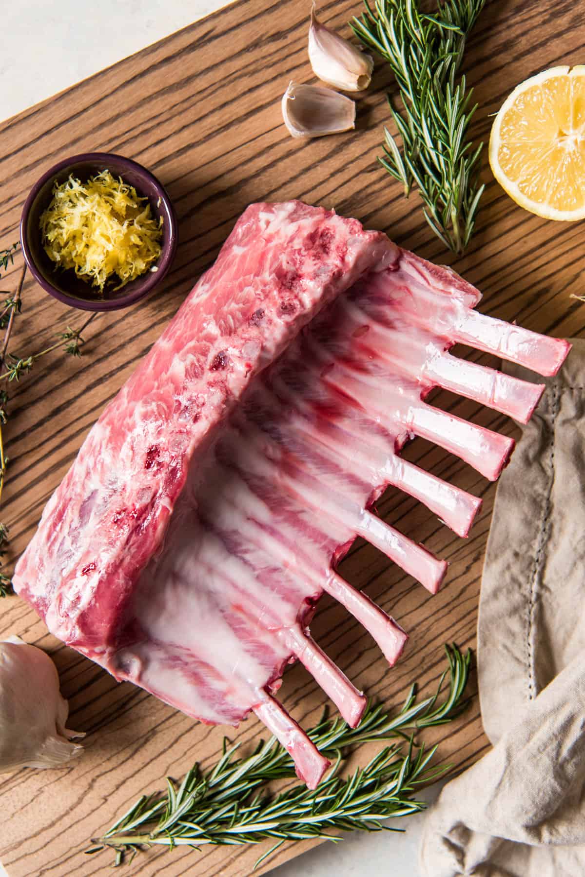 Raw rack of lamb sitting on a cutting board with other ingredients like lemon zest, garlic cloves and rosemary sprigs and ready to be prepared for roasting in the oven by first marinating.
