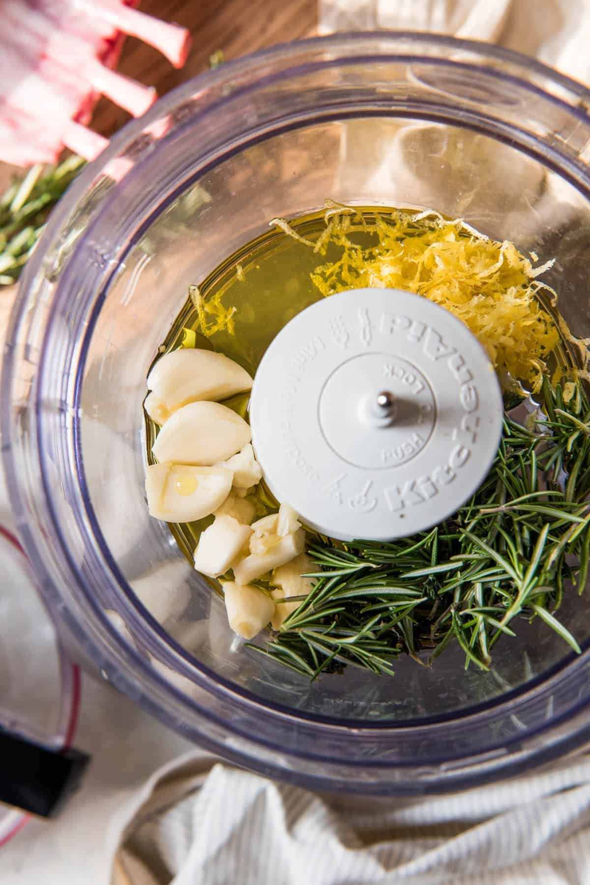Ingredients in a food processor ready to be blended into a rack of lamb marinade consisting of olive oil, garlic cloves, fresh rosemary and thyme, and the zest of one lemon.